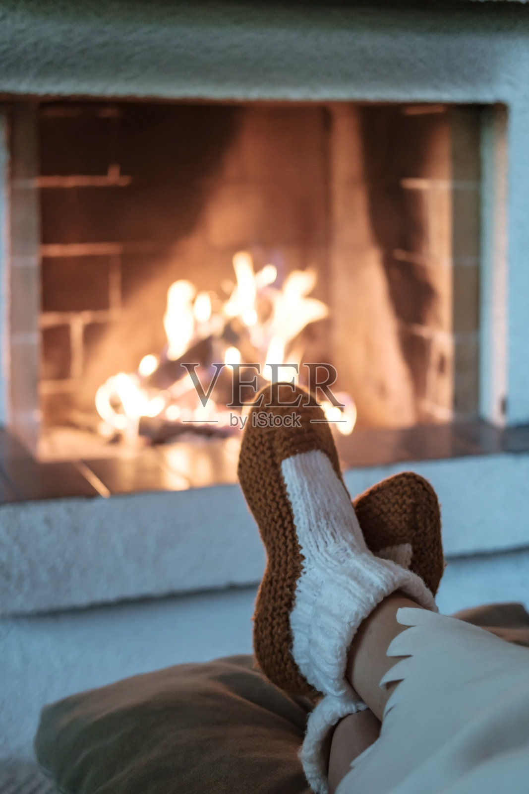 女性的腿在针织袜子覆盖着白色的毯子在模糊的背景开放的壁炉与点燃的柴火。冬天的晚上在家里放松，温暖和舒适。照片摄影图片