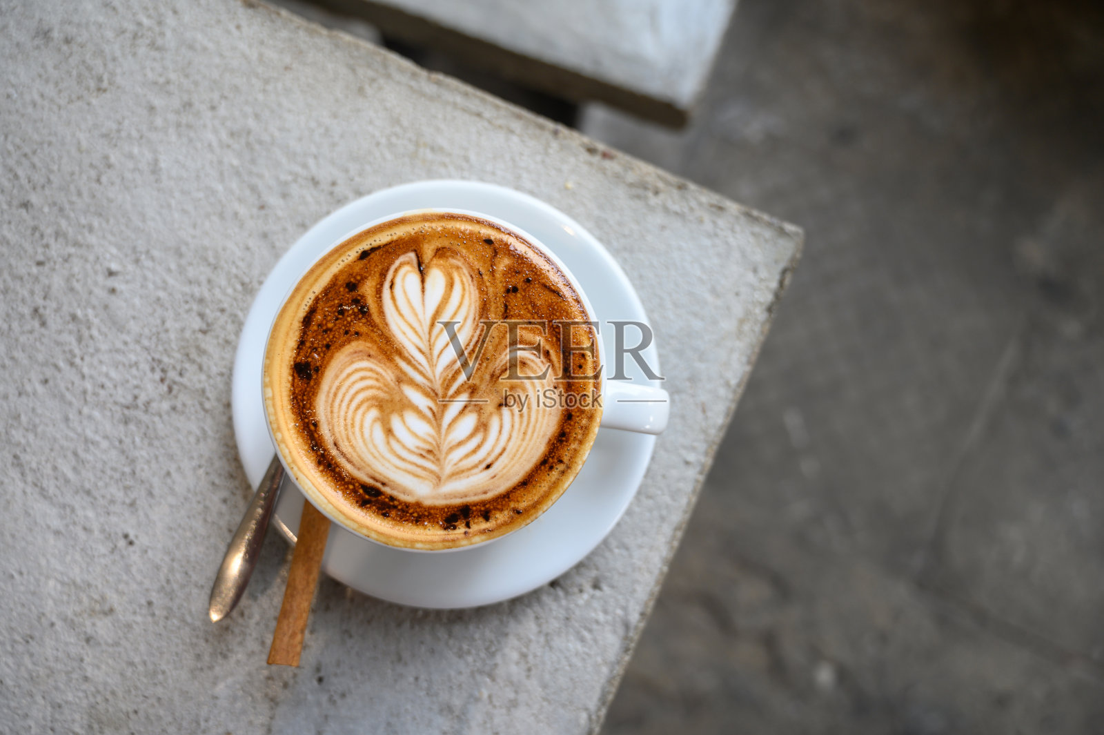 热卡布奇诺咖啡在白色陶瓷杯与肉桂和勺子在杯垫，放置在咖啡馆的水泥桌子的角落照片摄影图片