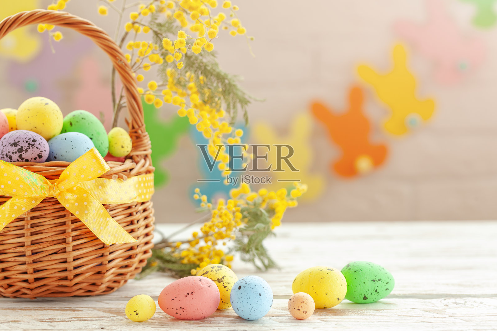 五彩缤纷的复活节彩蛋放在篮子里，以白色的木质为背景，用含羞草的花枝装饰。照片摄影图片