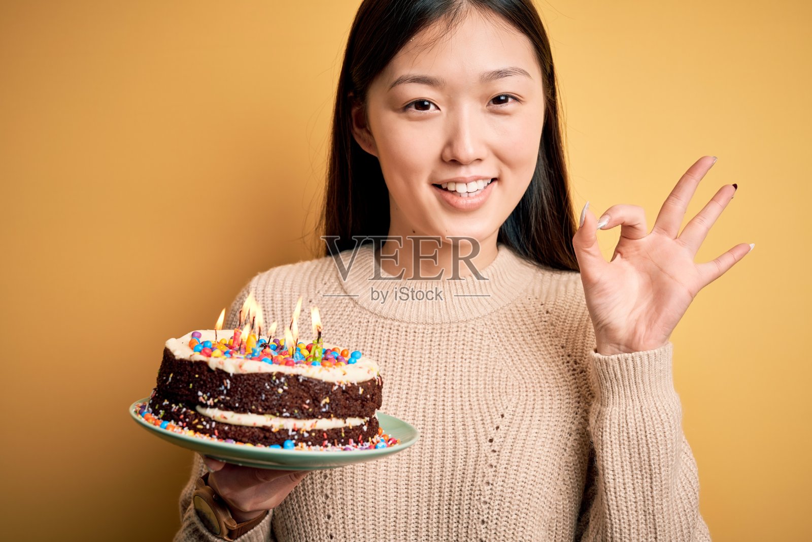 年轻的亚洲女人拿着生日蛋糕与蜡烛燃烧在黄色孤立的背景做ok手势与手指，优秀的象征照片摄影图片