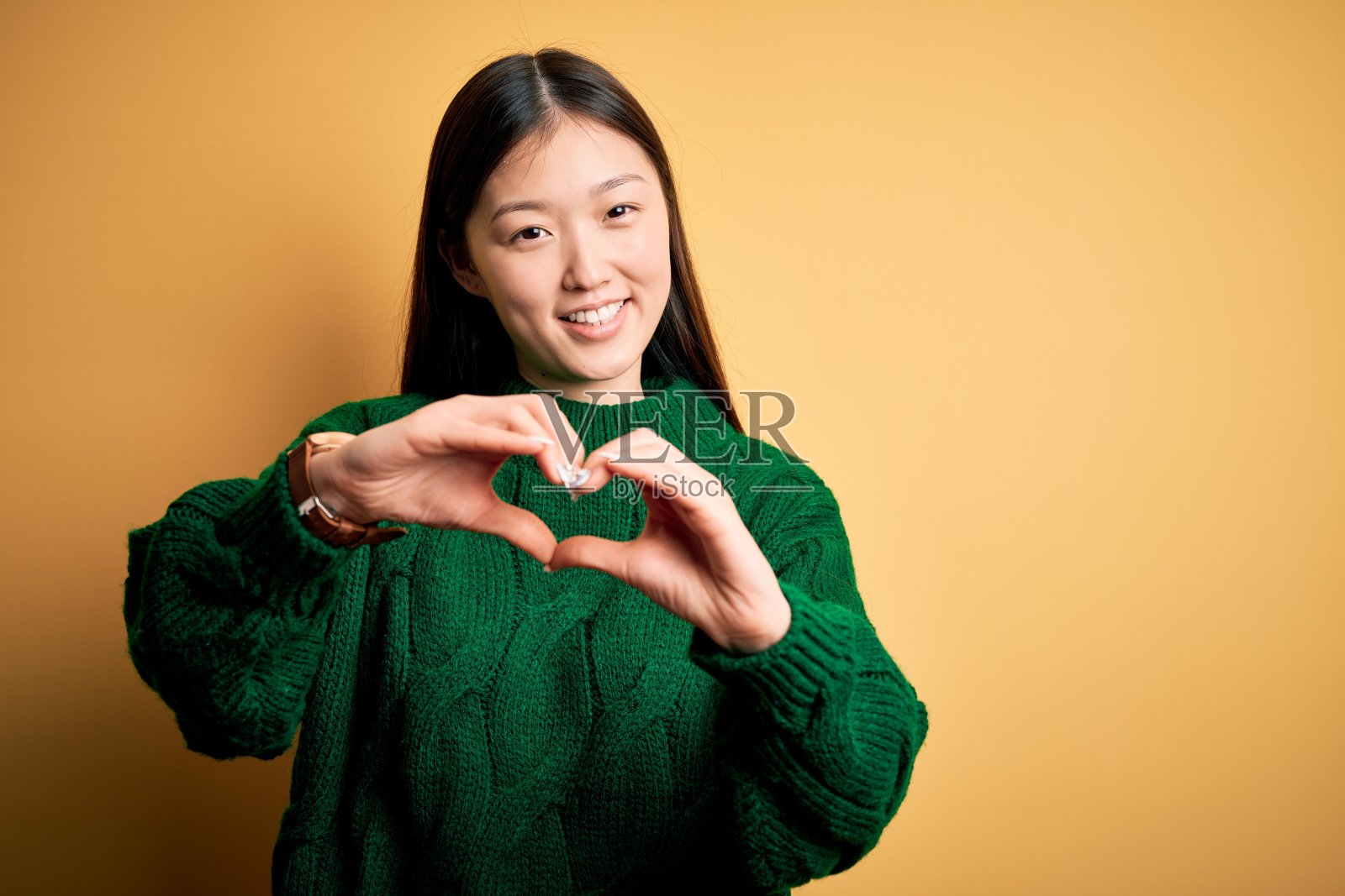 年轻美丽的亚洲女人穿着绿色的冬季毛衣在黄色孤立的背景微笑在爱做心形符号形状的手。浪漫的概念。照片摄影图片