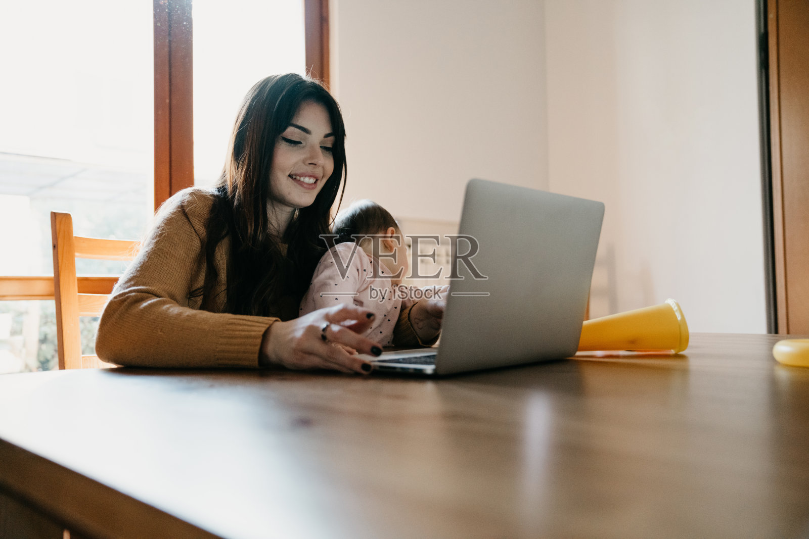 一位年轻的母亲在家里用电脑工作，同时照顾她的女儿照片摄影图片