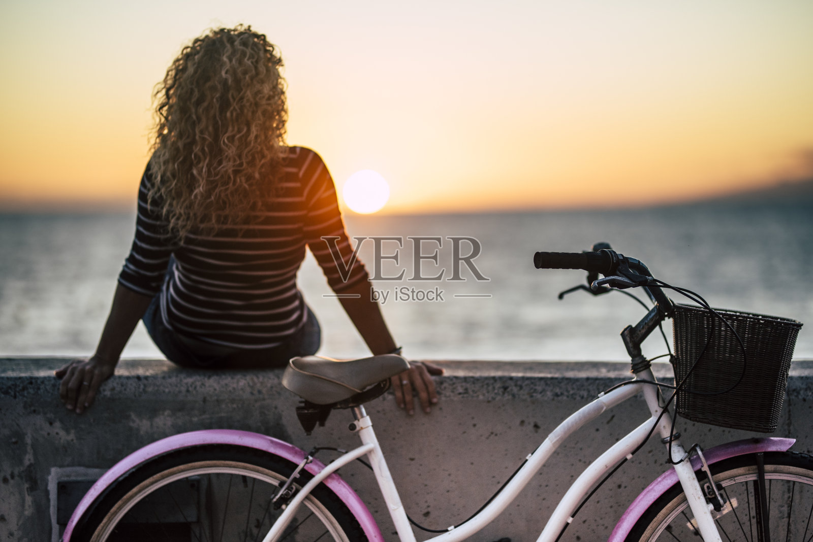 人们享受户外休闲活动，看着美丽的彩色日落，女人长长的卷发从后面看，享受自行车旅行后的放松-地平线和太阳与海洋照片摄影图片