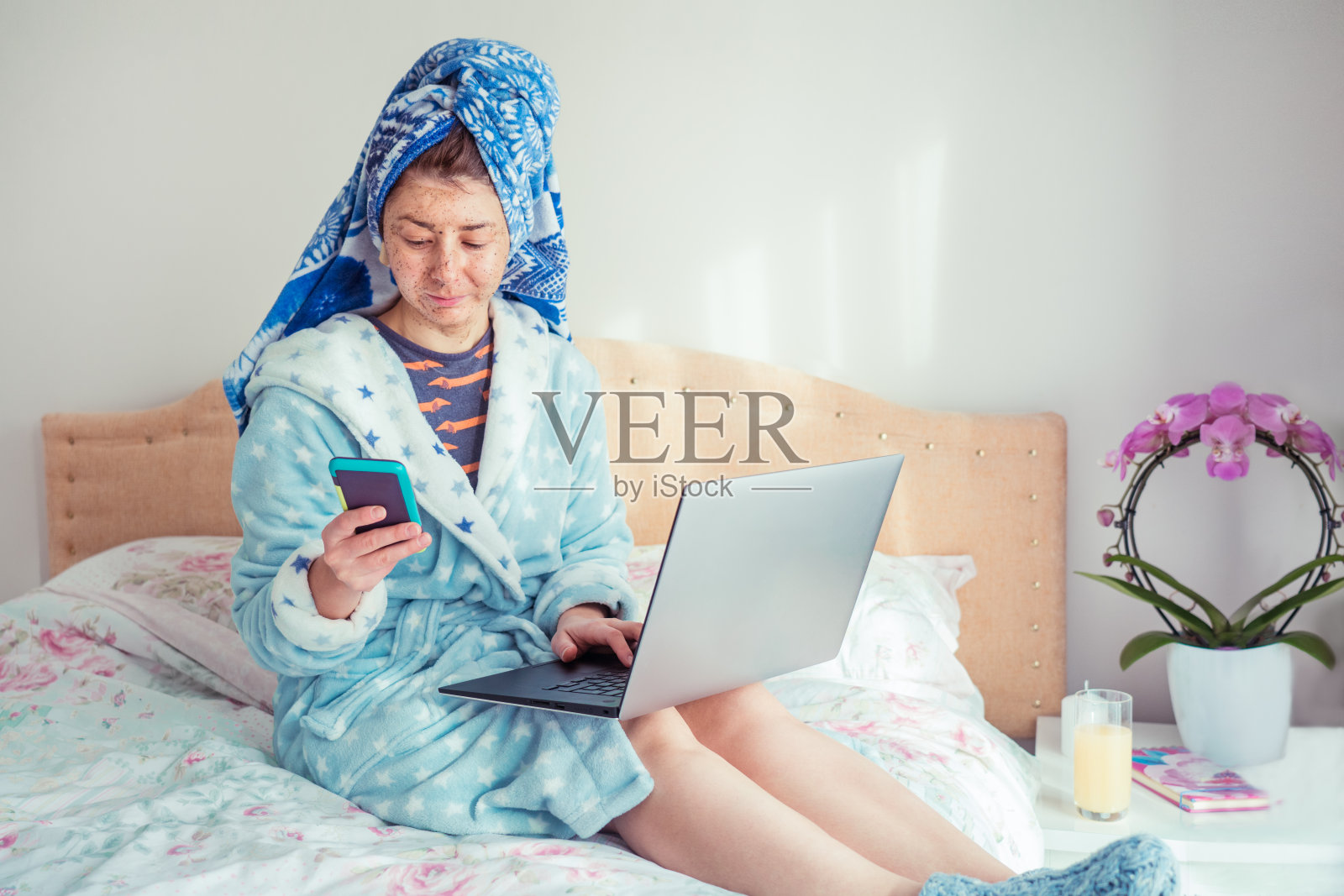 一个穿着浴袍，头戴毛巾的女人的肖像，在她的脸上敷着搓洗面膜，同时使用智能手机和笔记本电脑，坐在床上。在家工作。远程工作。照片摄影图片