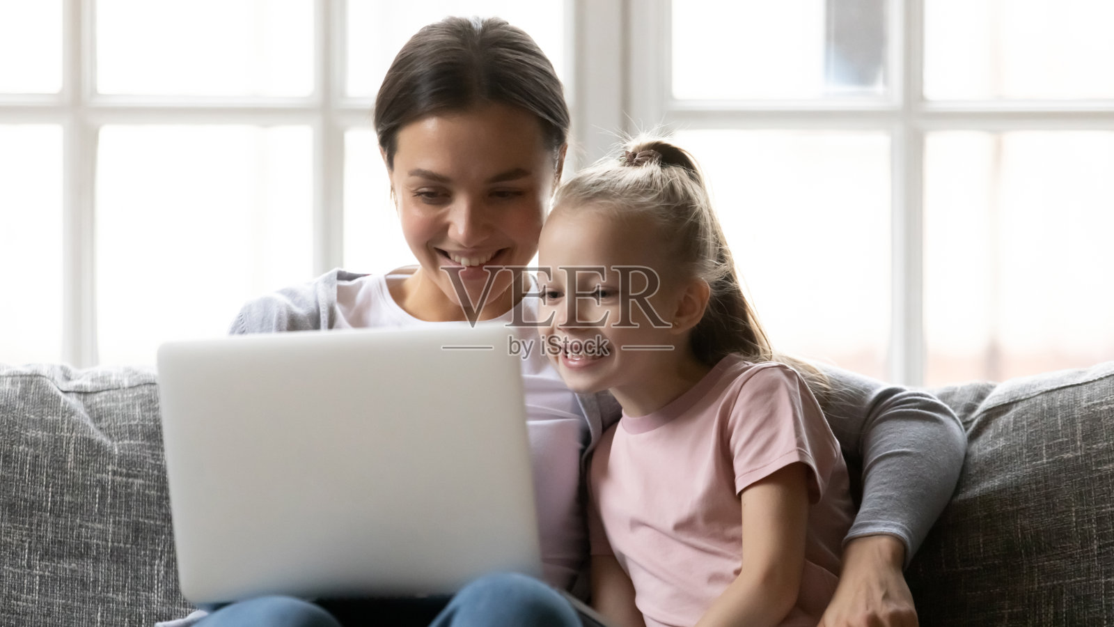 年轻微笑的母亲看有趣的视频在笔记本电脑和女儿。照片摄影图片