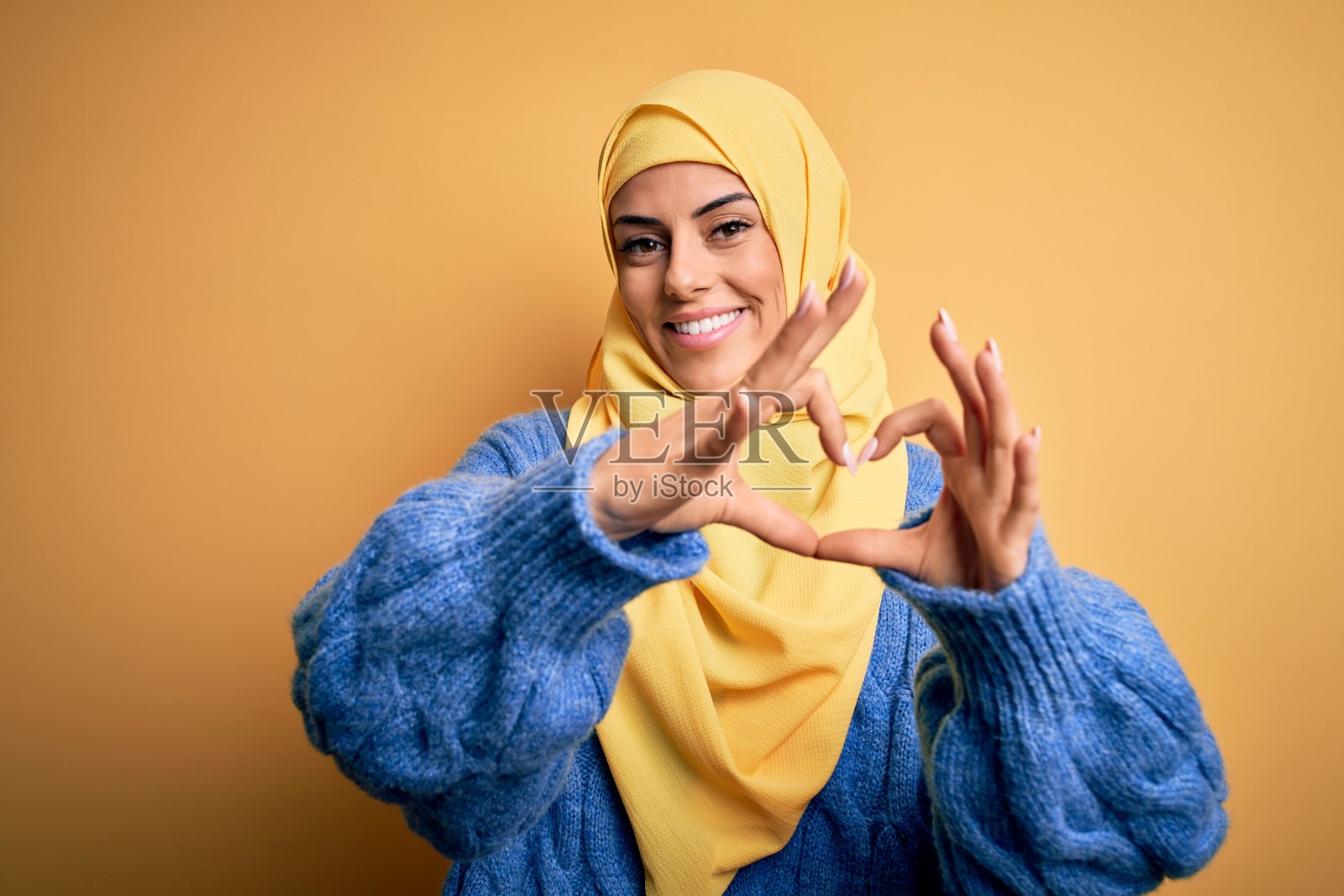 年轻美丽的黑发穆斯林妇女戴着阿拉伯头巾在孤立的黄色背景微笑在做心形符号形状的手。浪漫的概念。照片摄影图片