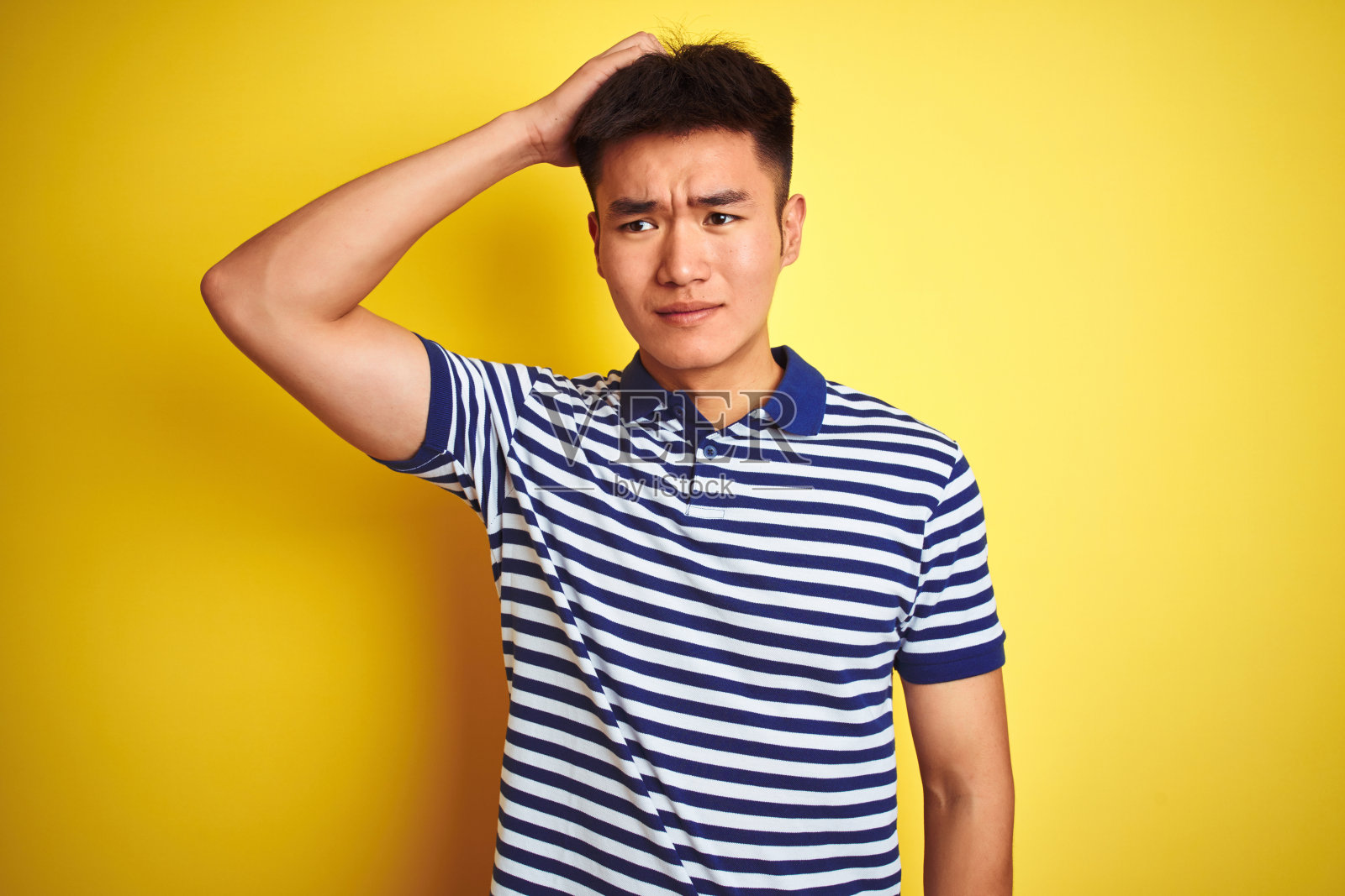 年轻的亚洲华人男子穿着条纹马球站在孤立的黄色背景困惑和疑惑的问题。不确定用怀疑，用手思考。沉思的概念。照片摄影图片