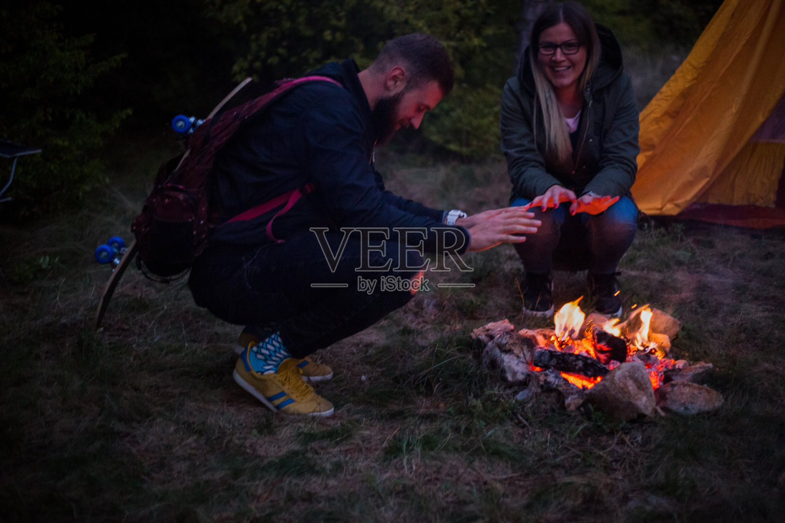 漂亮的一对，围坐在篝火旁。背景是一个帐篷，那人用一根棍子弹火。照片摄影图片