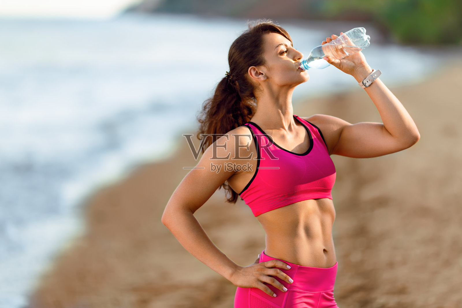 年轻的女运动员在海滩上喝水。照片摄影图片