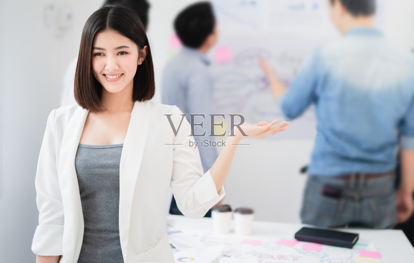 年轻的亚洲女企业家一边自信地微笑着，一边举手向欢迎或介绍会议，背景与同事在会议董事会讨论，分析商业计划的概念照片摄影图片