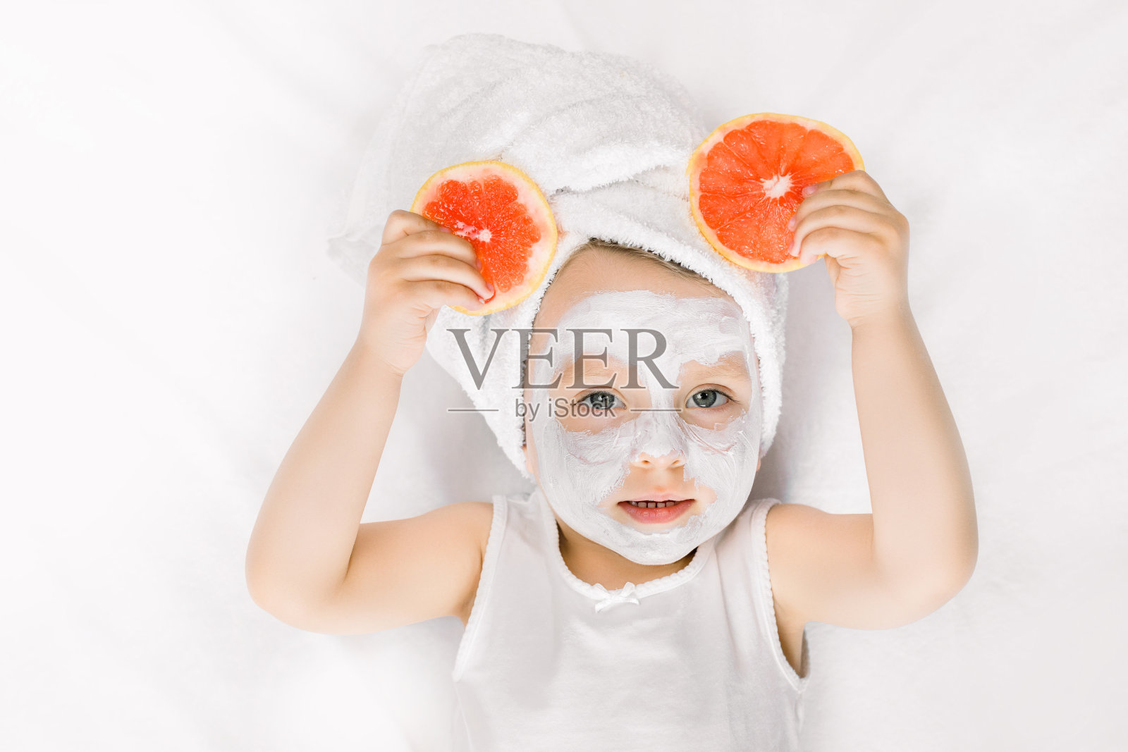 美容、水疗、儿童卫生理念。可爱的小女孩，头上裹着毛巾，脸上戴着白色的粘土面具，对着镜头摆姿势，手里拿着葡萄柚片照片摄影图片