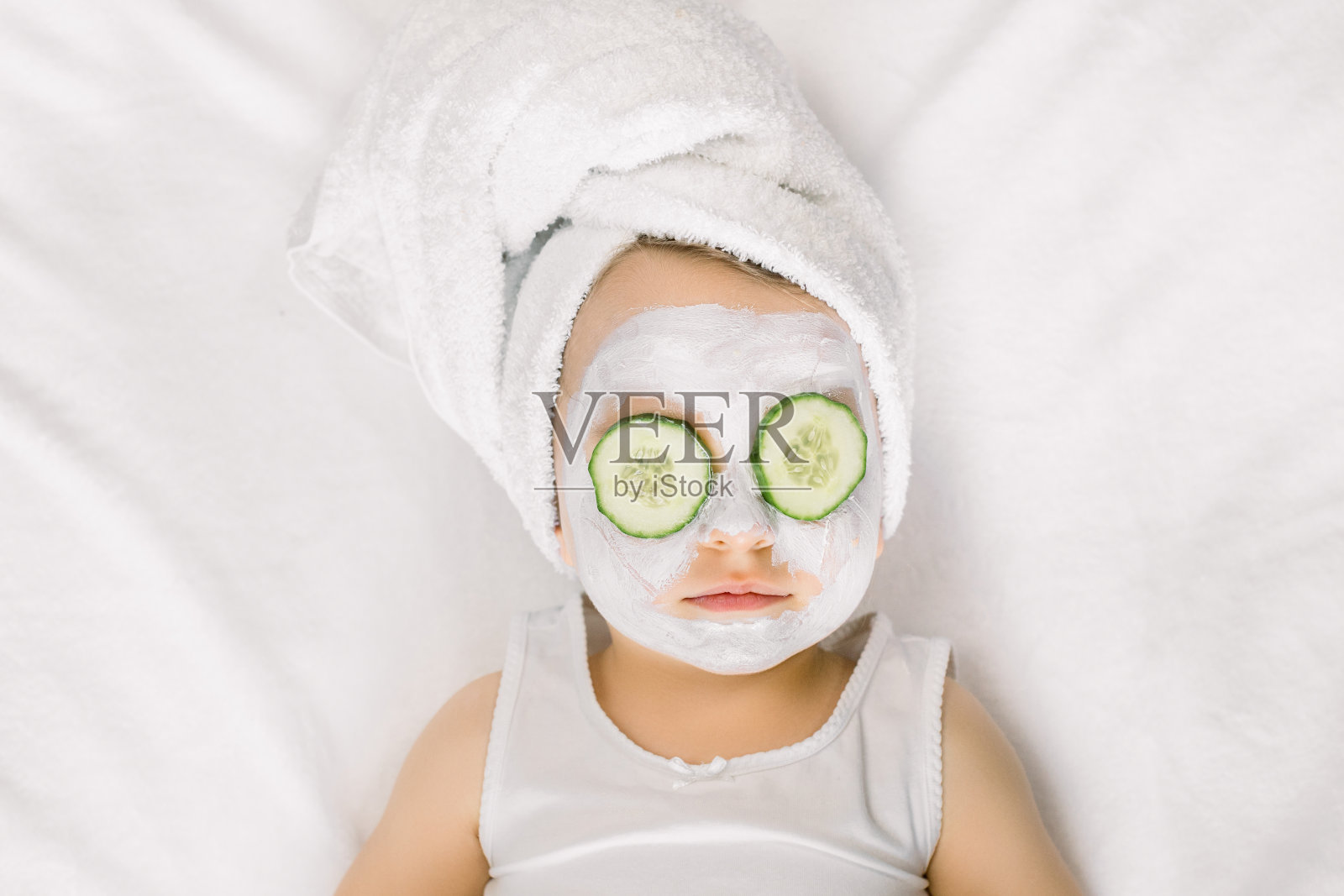 一个可爱的小女孩在白色的浴巾和黄瓜在她的眼睛躺在白色的背景。宝宝用毛巾包裹，用面膜和黄瓜片敷在眼睛上照片摄影图片