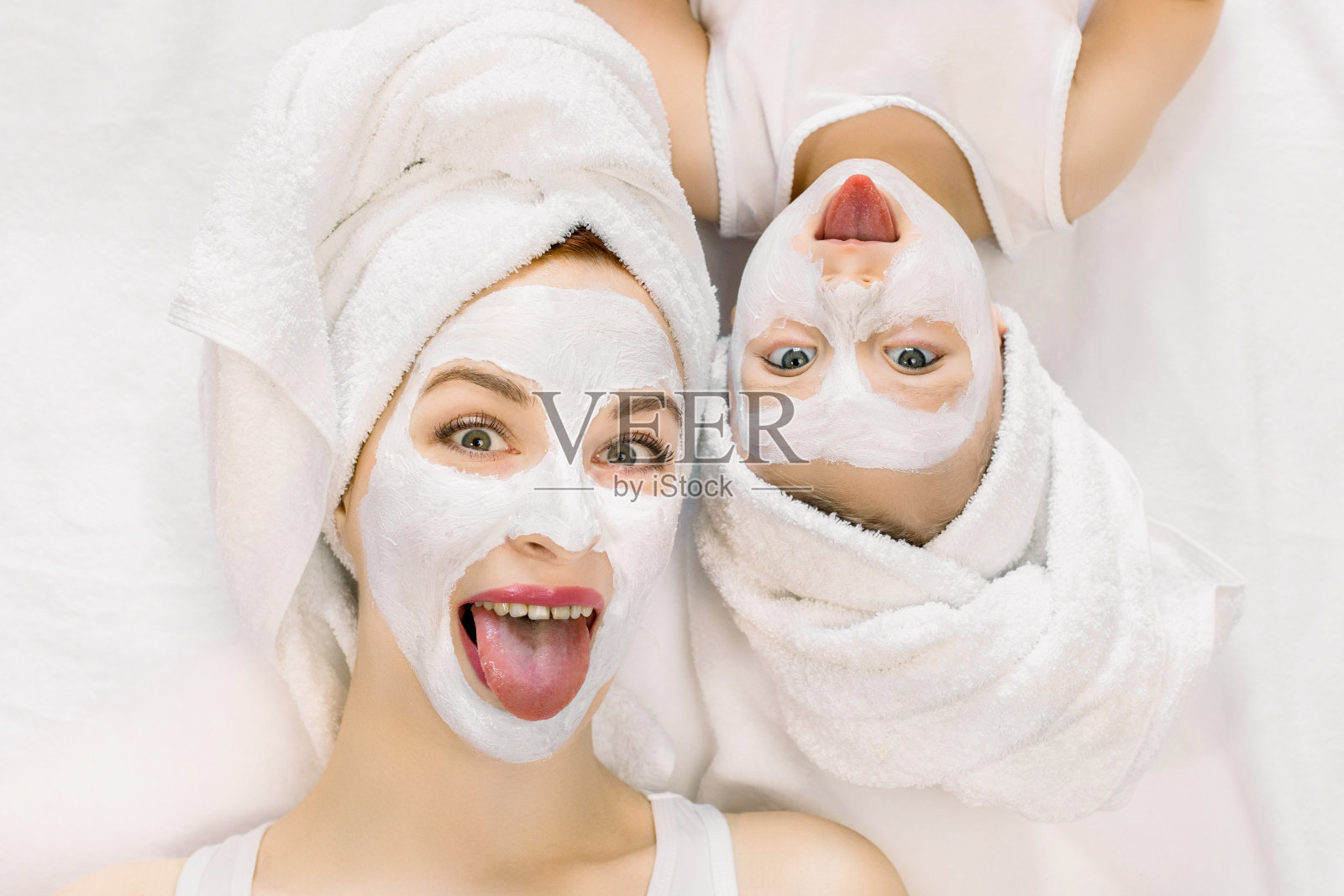 这是一张有趣的照片，快乐的妈妈和小宝贝女儿展示他们的舌头，头发裹在浴巾里，躺在家里的床上，做一个粘土面膜。家庭卫生间护肤照片摄影图片