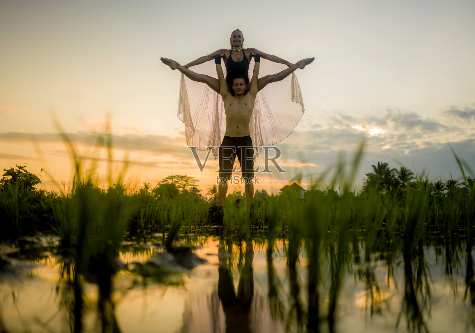 户外日落acroyoga锻炼-年轻快乐和健康的夫妇练习跨越瑜伽练习在美丽的稻田享受自然和健康的生活方式做杂技姿势照片摄影图片