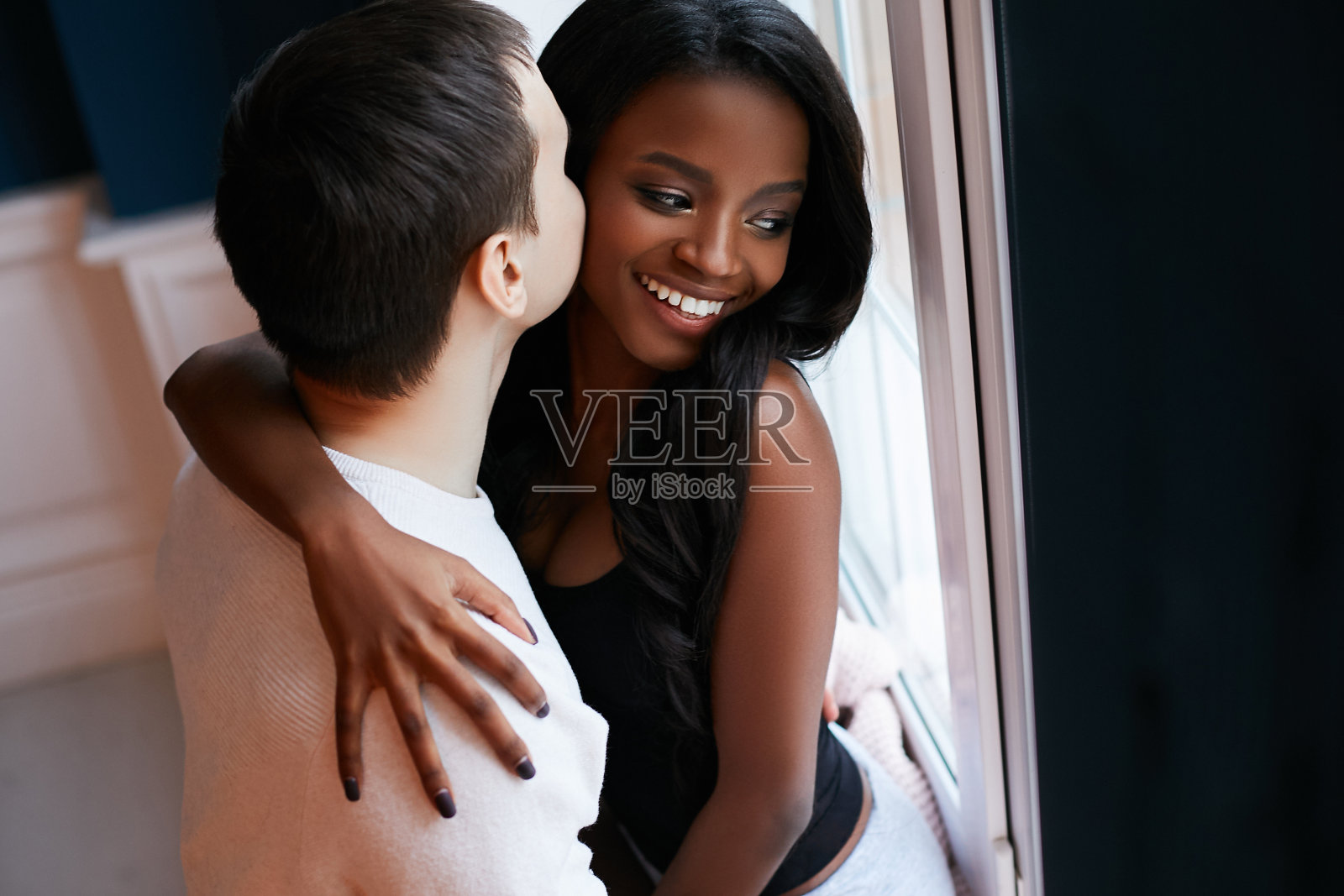 幸福、相爱的年轻异族情侣站在窗边拥抱。美丽的黑人女孩微笑着亲吻。照片摄影图片
