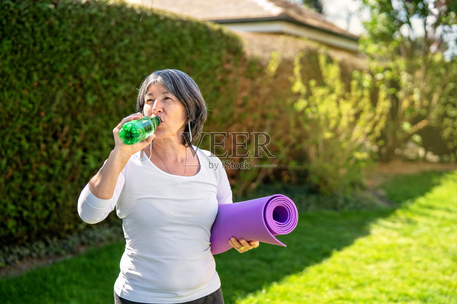 成熟的成年女性戴着耳机和健身手表，在户外运动后喝着水，拿着瑜伽垫看着镜头。老年人健康的生活方式、活力、保健理念。副本的空间。照片摄影图片