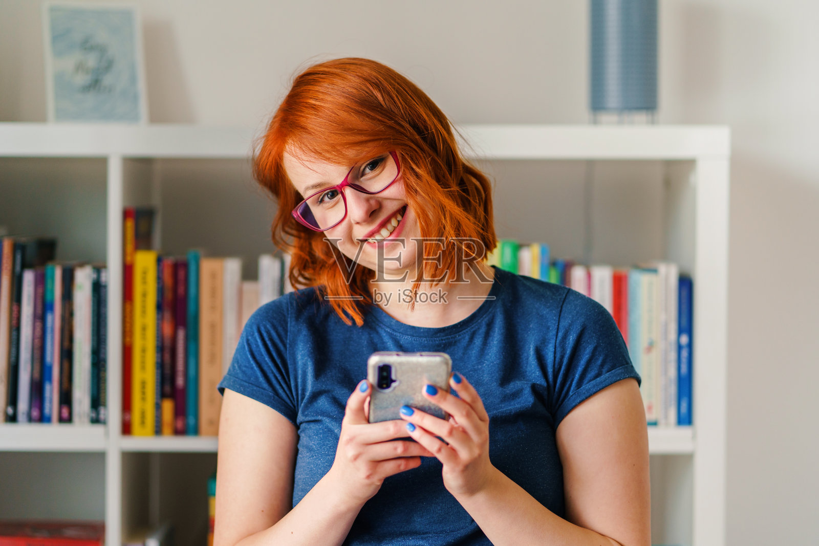 特写年轻的白人女子美丽的女性自然的美丽女孩橙色的头发和蓝色的t恤拿着手机在家里前面模糊的书架上戴眼镜微笑照片摄影图片