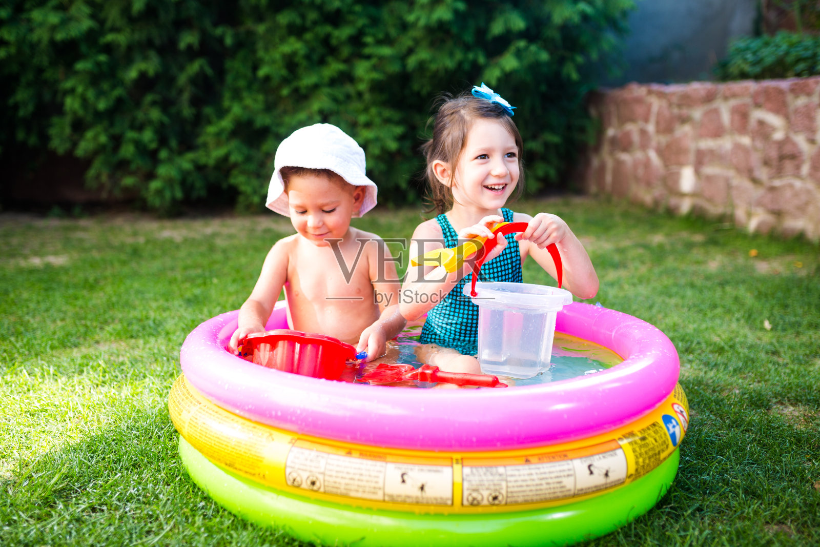 以院子里的儿童夏季游戏为主题。白种兄妹坐在水里玩塑料玩具桶，围着充气儿童水池。夏天很热，穿着泳衣休息照片摄影图片