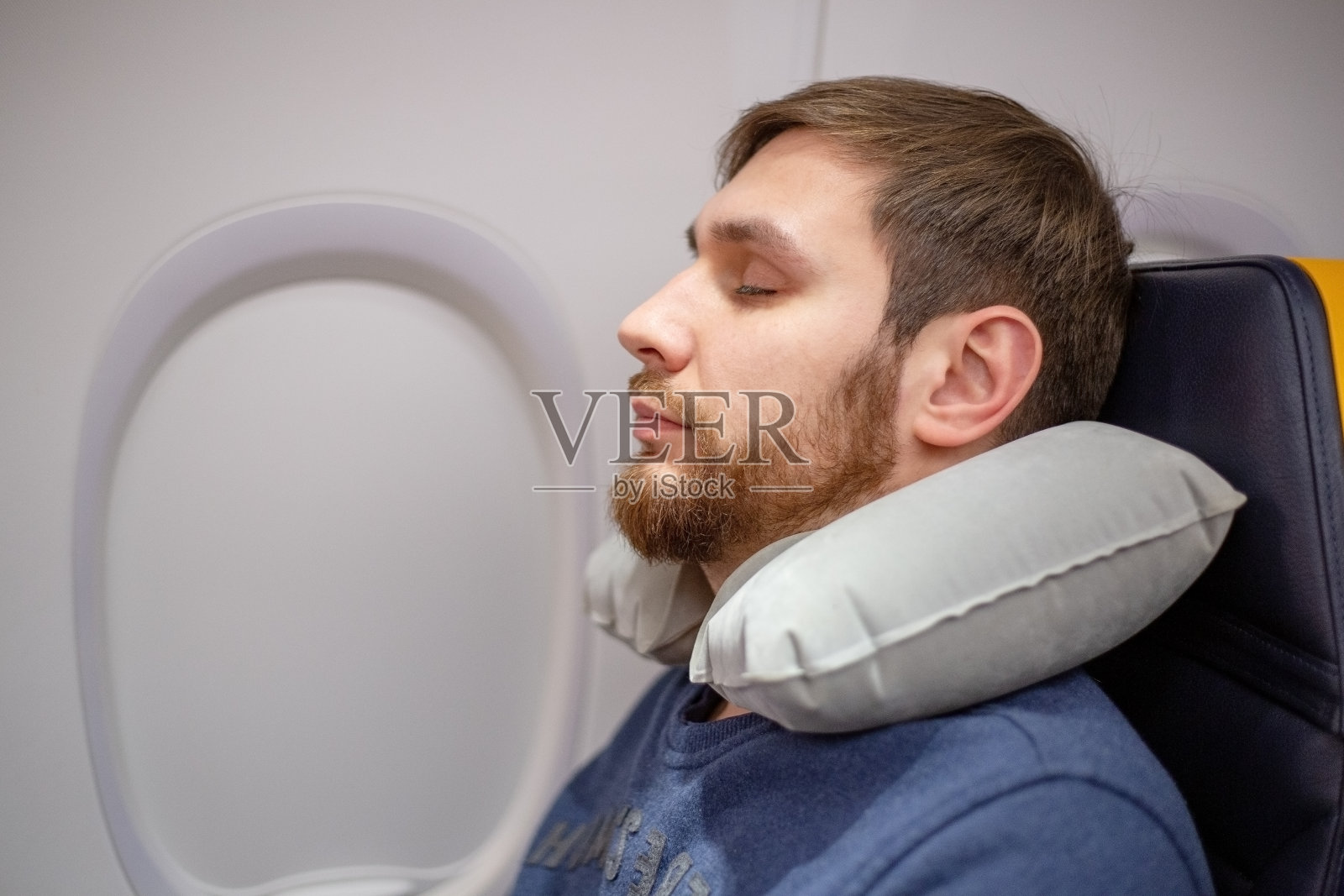 一名年轻帅气的欧洲男子30岁，留着胡子，在飞机上枕着充气枕睡觉。舒适，飞机上的压力，交通工具，旅行。股票的照片。照片摄影图片