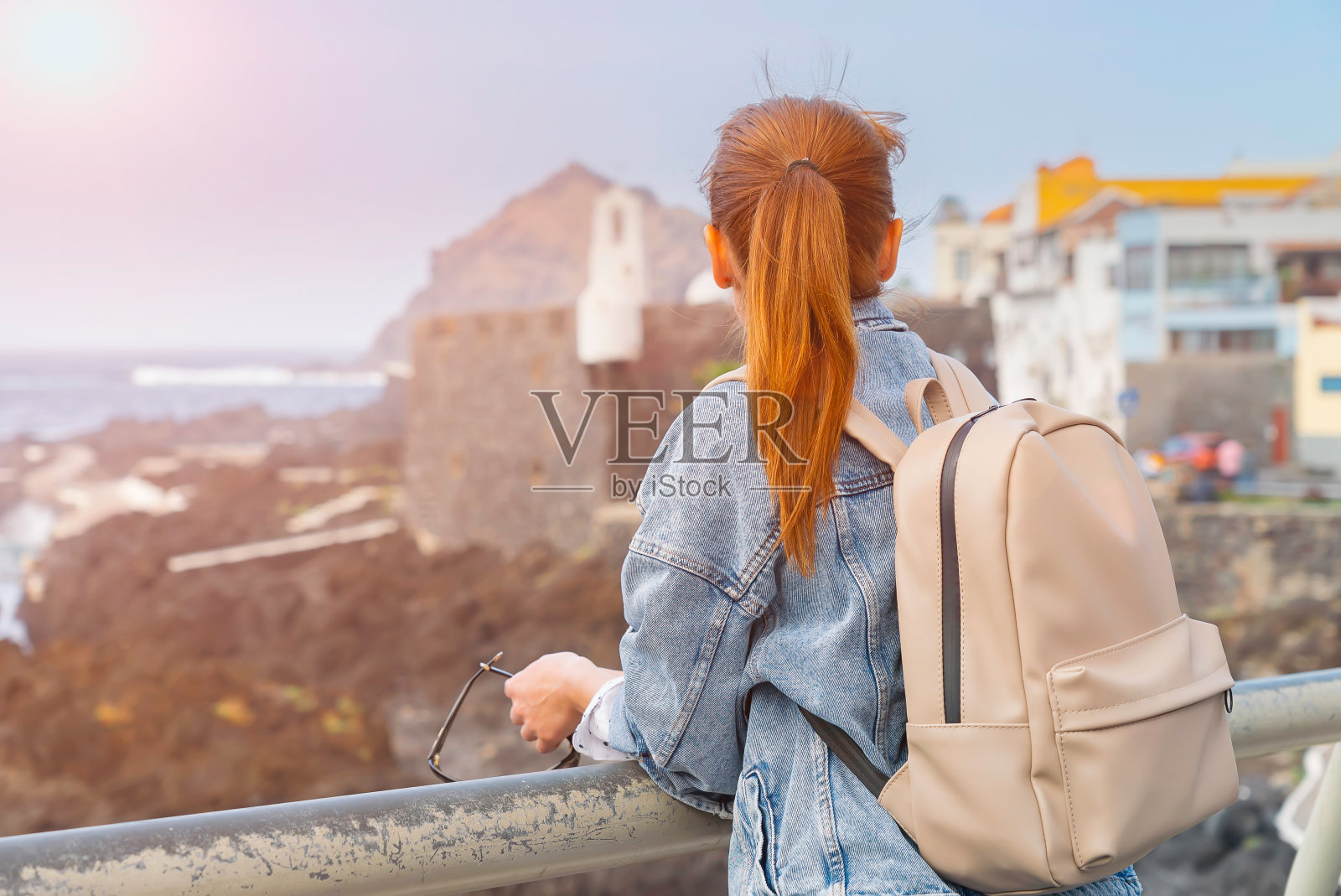 在一个阳光明媚的日子里，一个背着背包的红发女孩在欣赏特内里费岛Garachico的海洋和建筑。旅行的概念。女游客探索地点与一个小背包。照片摄影图片