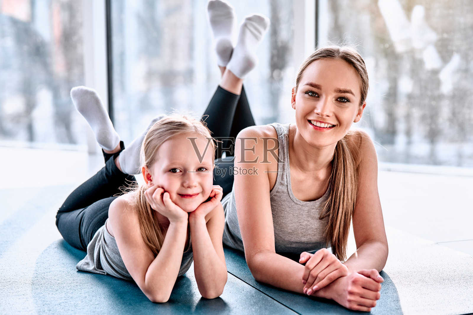 美丽的年轻女子和迷人的小女孩看着镜头和微笑，而躺在瑜伽垫在健身大厅照片摄影图片