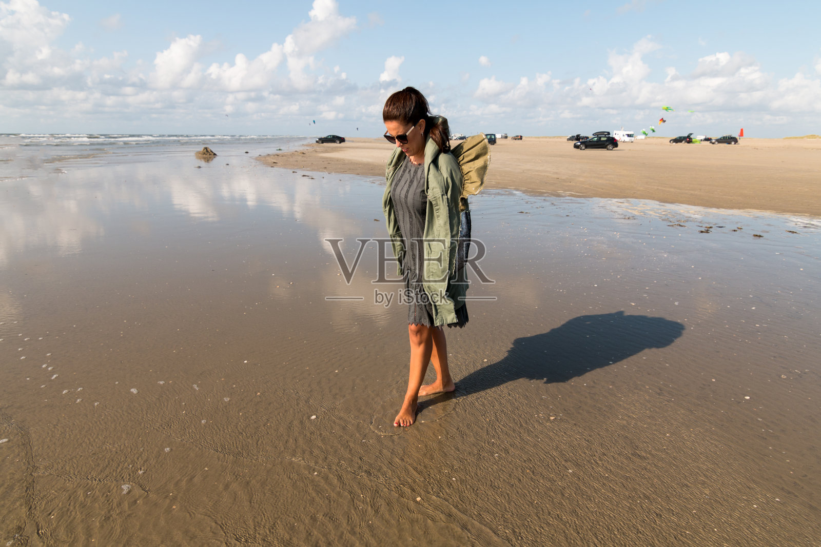 在丹麦的Romo海滩上，一名女子赤脚走在水里，她穿着一件绿色的外套，戴着太阳镜，沙滩上有一些汽车作为背景照片摄影图片