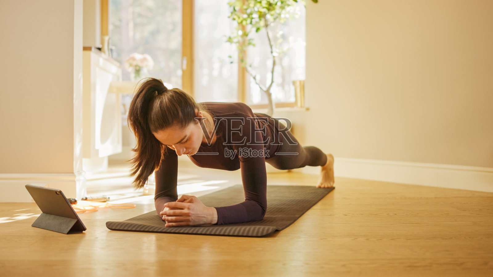 Strong Beautiful Fitness Girl in Workout Clothes Does Stretching Yoga Exercises at Home, Uses Digital Tablet for Online Exercise, Workout, Streaming Fitness。在明亮的客厅里进行家庭运动照片摄影图片