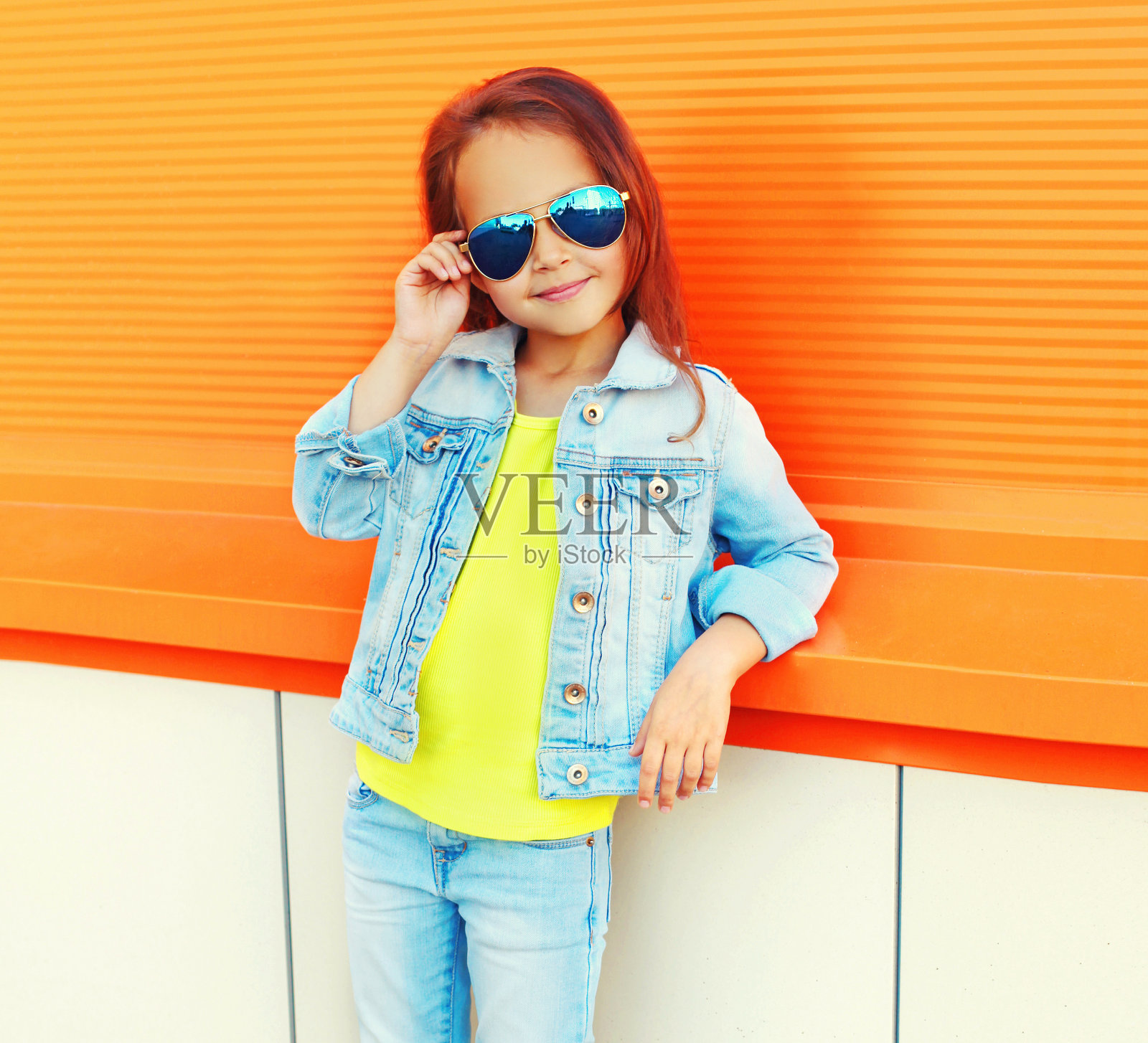 一个小女孩的肖像，戴着太阳镜，牛仔裤夹克在城市街道上橙色的背景照片摄影图片
