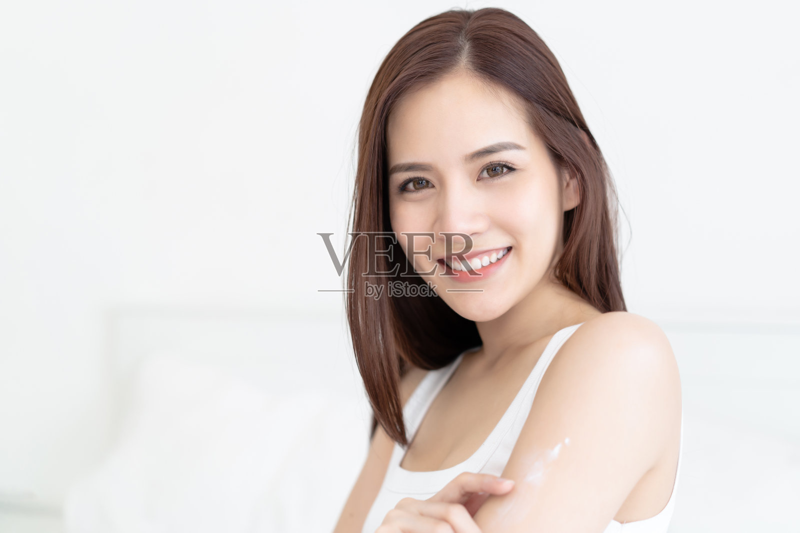 美丽的亚洲女人涂抹身体乳液在她的迷人的手臂在她的早上例行公事和看在卧室的相机。皮肤护理，美容，健康和健康理念照片摄影图片