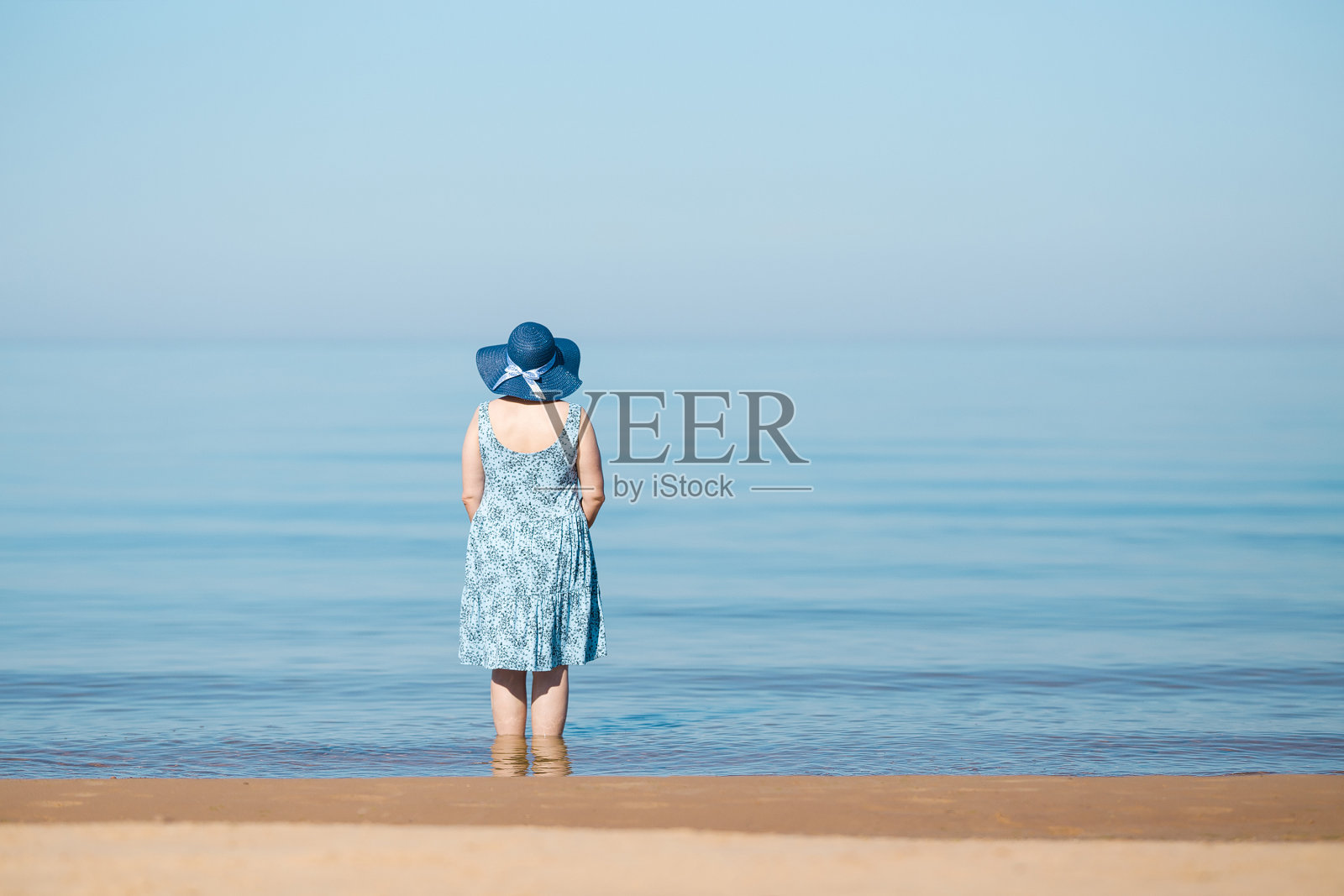 成熟的女人穿着衣服，戴着草帽，独自站在水中。后视图。在温暖的夏日早晨，平静的大海和浅蓝色的天空。空的地方，情感，鼓舞人心的文本，引用或语录。照片摄影图片