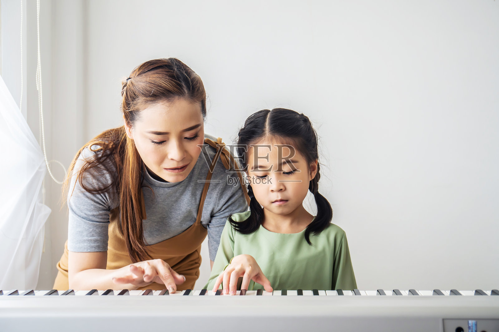 快乐的小亚洲女儿弹钢琴与母亲在家里，母亲教女儿弹钢琴，他们弹琴唱歌。他们玩得很开心。照片摄影图片