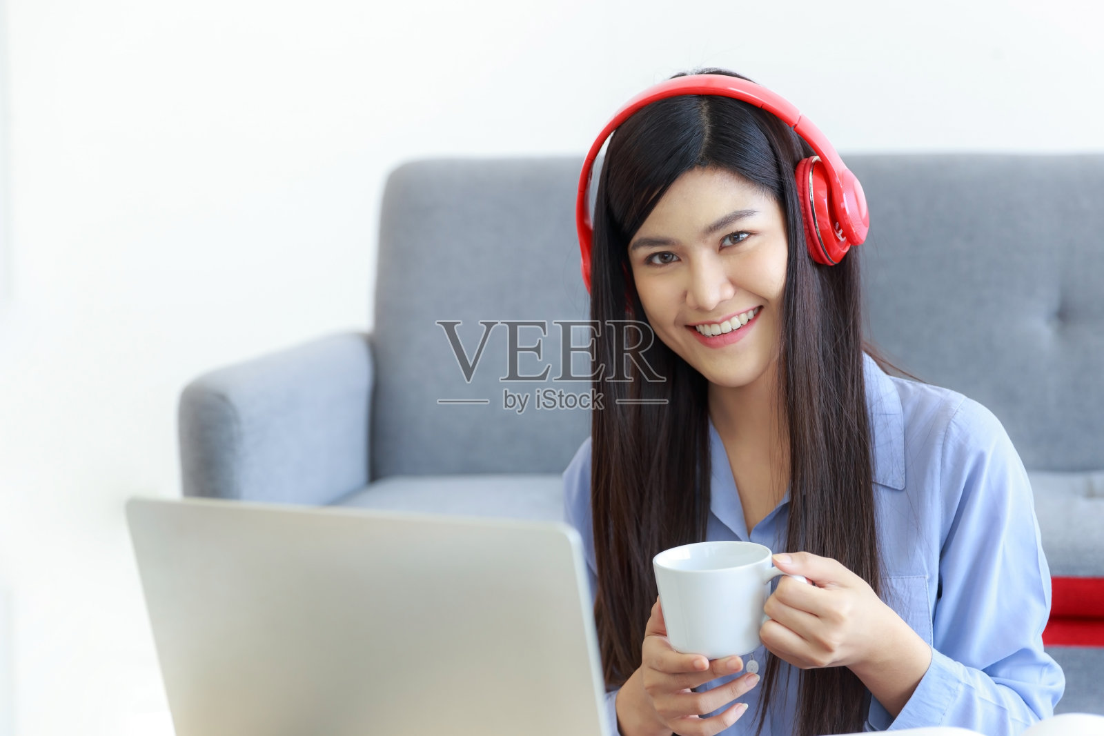 一位面带微笑的亚洲妇女手里拿着一个咖啡杯，在家里的客厅里享受着一台笔记本电脑。照片摄影图片