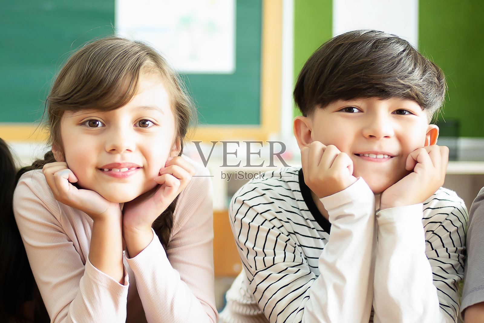 回学校!快乐的小学生坐在课桌上，手托着下巴，看着教室里的相机。教育，小学，童年观念。照片摄影图片
