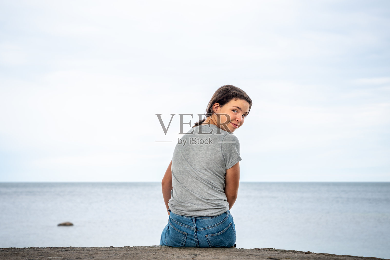 年轻的女孩坐在悬崖上向后看，以水平线为背景。照片摄影图片