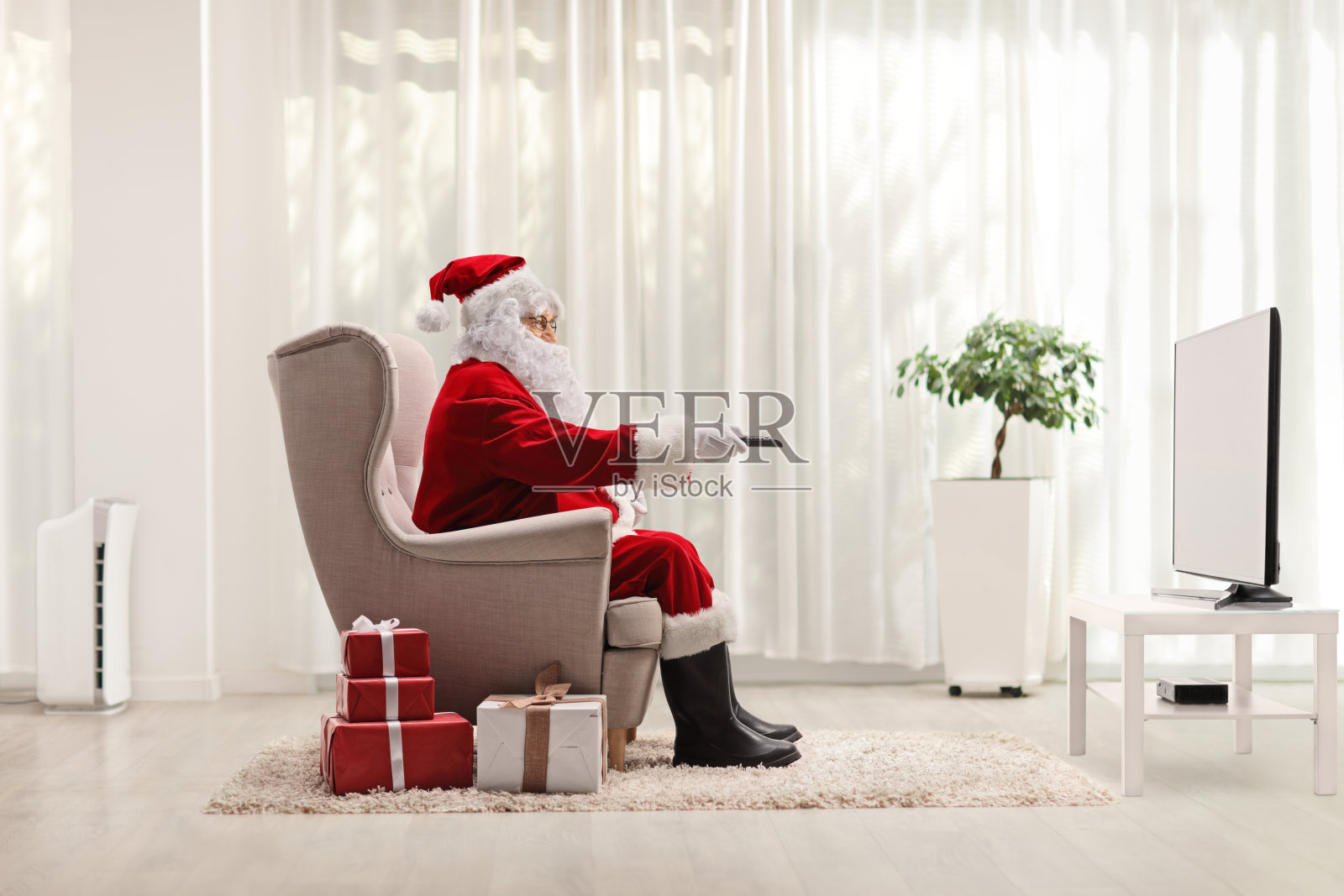 圣诞老人坐在扶手椅上，遥控器放在电视机前，家里的地板上放着礼物照片摄影图片