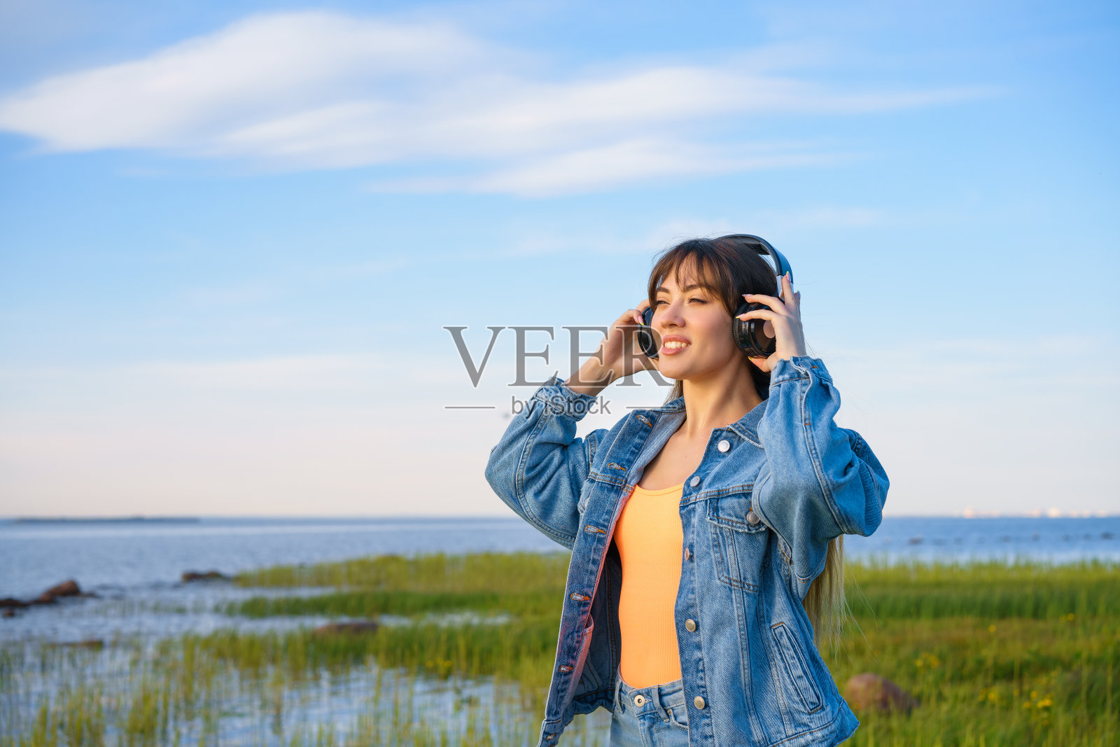 一个穿着牛仔夹克的漂亮女孩在蓝天和大海的背景下摆姿势，用无线耳机听着音乐照片摄影图片