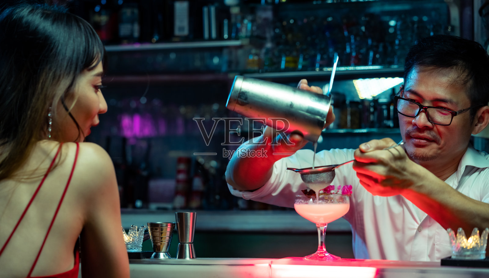 职业男调酒师准备混合酒精鸡尾酒服务给坐在酒吧柜台的女人。亚洲调酒师，将混合烈酒倒入装饰性的鸡尾酒杯中。照片摄影图片