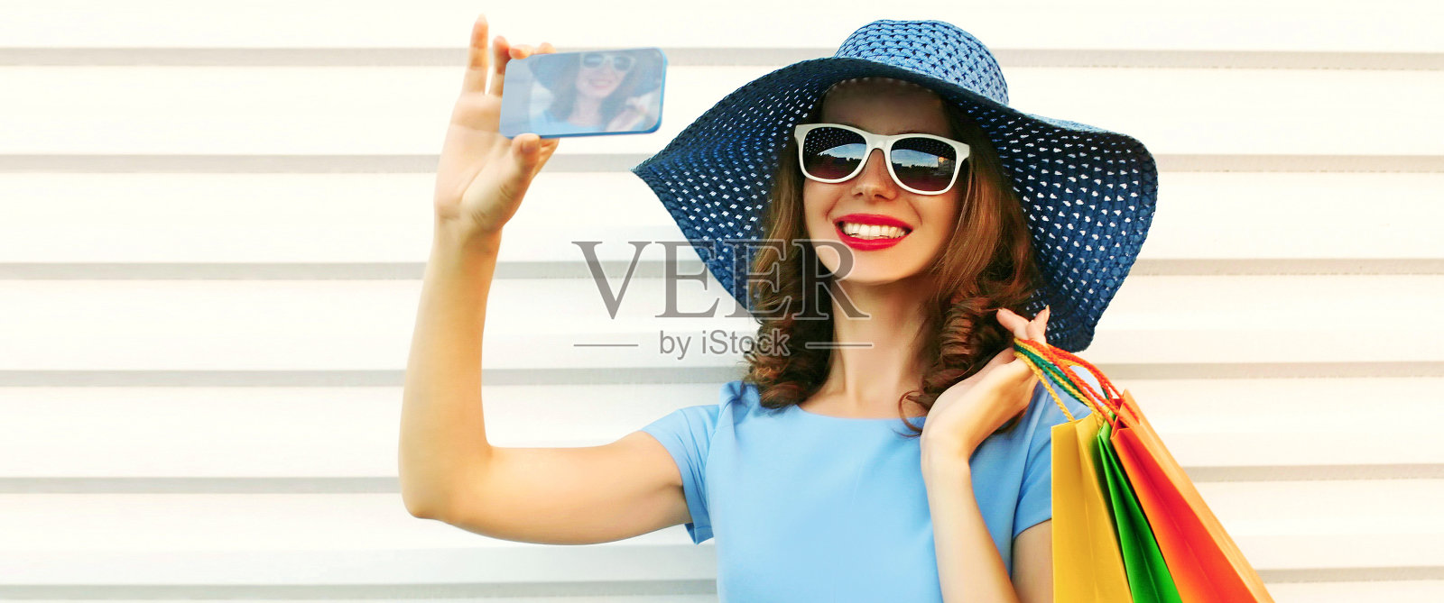 照片近距离微笑的年轻女子用智能手机自拍与购物袋戴着蓝色的夏季草帽在白色背景照片摄影图片