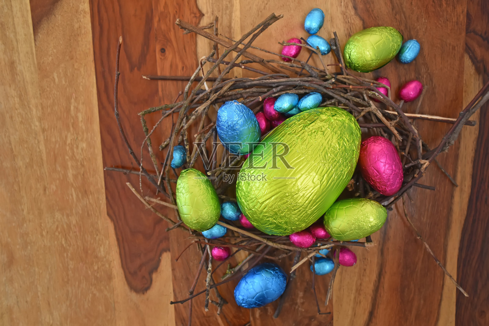 用锡纸把五颜六色的复活节彩蛋包裹起来，有粉色、绿色、蓝色和黄色，放在用树枝和小树枝做成的天然巢里。中间有一个大鸡蛋，它是在多粒棕色木制背景下。照片摄影图片