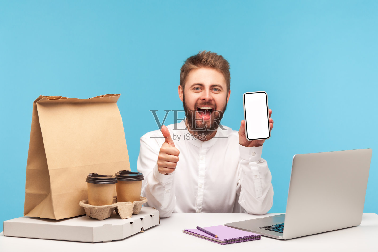 快乐满意的男性客户举起大拇指举着白色屏幕的智能手机，咖啡披萨和纸袋躺在桌子上，在线订购和快速送货照片摄影图片