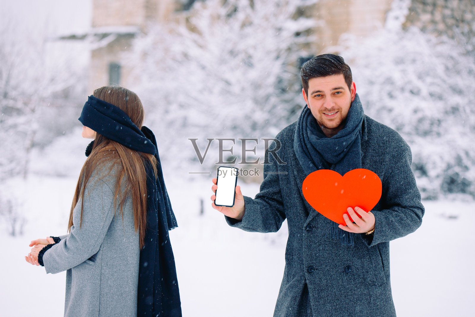 年轻浪漫的情侣，惊喜户外，冬天。男人拿着红心和手机与空白空白屏幕模拟拷贝空间看着相机。闭着眼睛的盲人女人。照片摄影图片