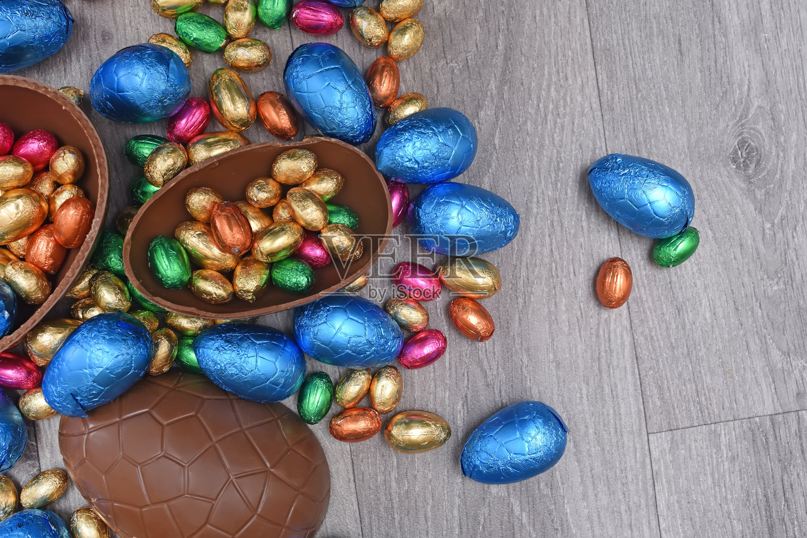 一堆或一组多颜色和不同大小的彩色锡箔包装的巧克力复活节彩蛋，粉红色，蓝色，金色和绿色。一个大的半块棕色牛奶巧克力鸡蛋，里面有迷你的鸡蛋，在灰色的木头背景上。照片摄影图片
