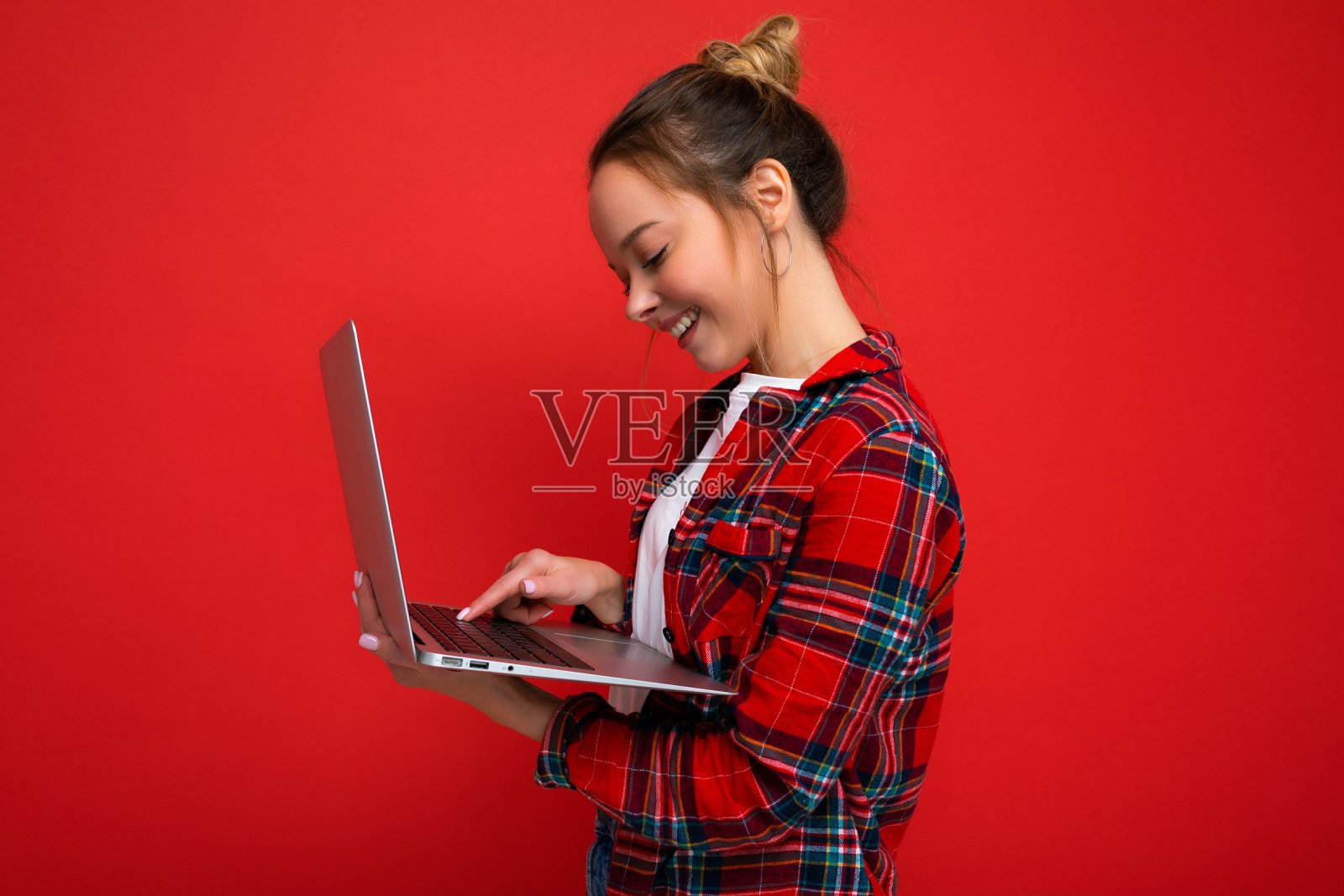 侧面照片的漂亮微笑年轻的女人拿着上网本看屏幕在键盘上打字穿着红色衬衫孤立在红色的墙背景照片摄影图片