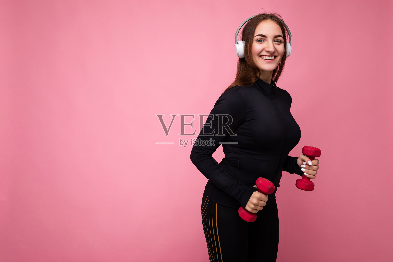 有吸引力的积极微笑的年轻黑发女人穿着黑色运动服孤立在粉红色的背景墙上做健身使用哑铃戴白色蓝牙耳机听好音乐和有趣的看着相机照片摄影图片