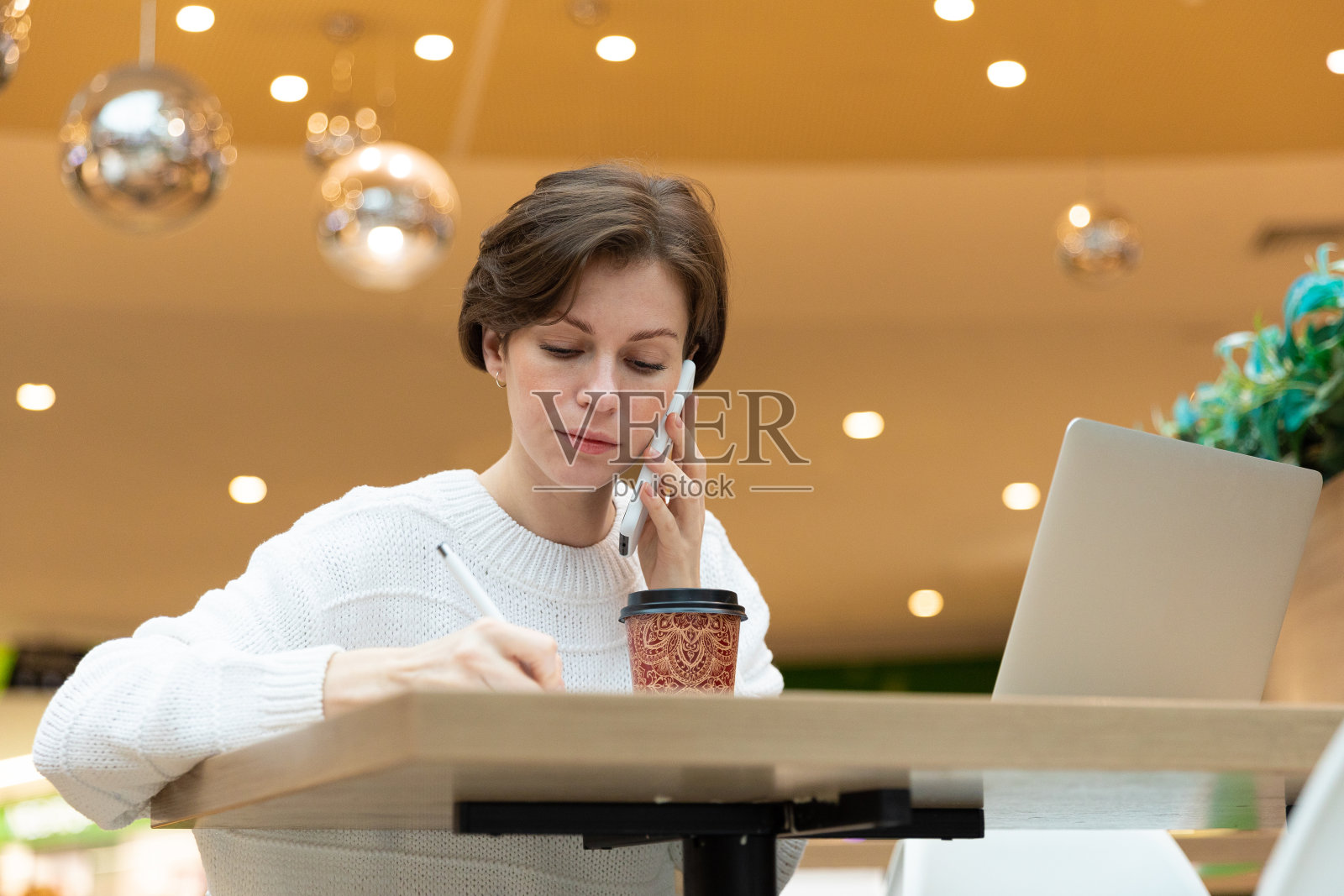 程序员坐在咖啡馆里的一台笔记本电脑前，在记事本上写着下面的照片。穿白毛衣的黑发女人，自由职业者的概念照片摄影图片
