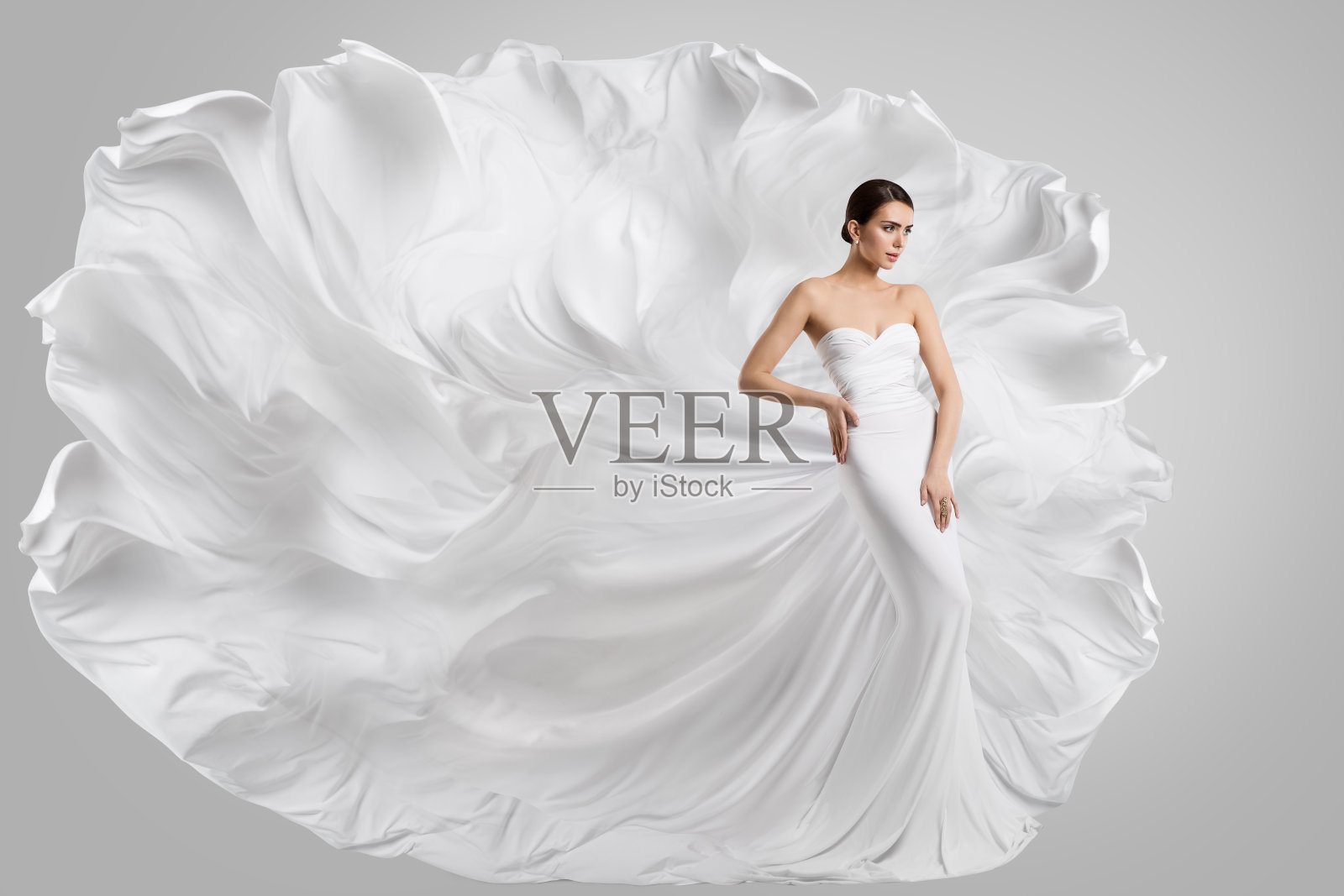 穿着白色婚纱的女人。时尚新娘模特在风中飘扬的长礼服。灰色的背景。侧视图照片摄影图片
