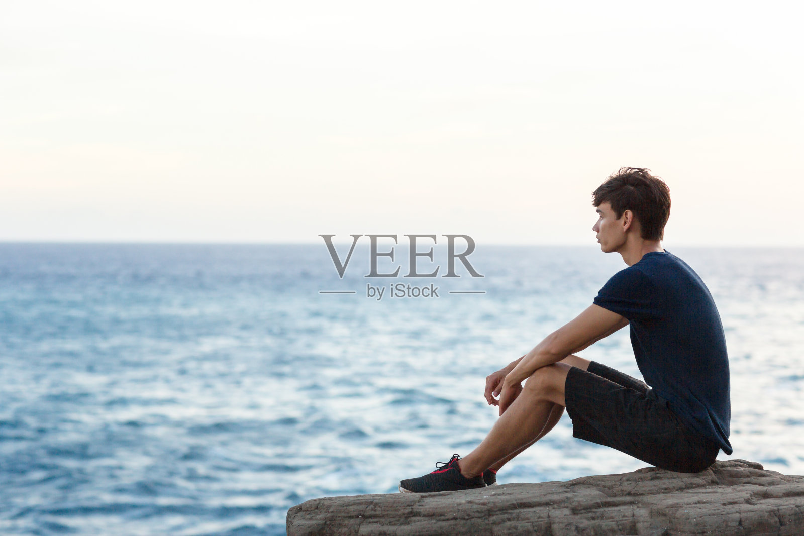 一个轻松的年轻人独自坐在悬崖上看海边的景色。照片摄影图片