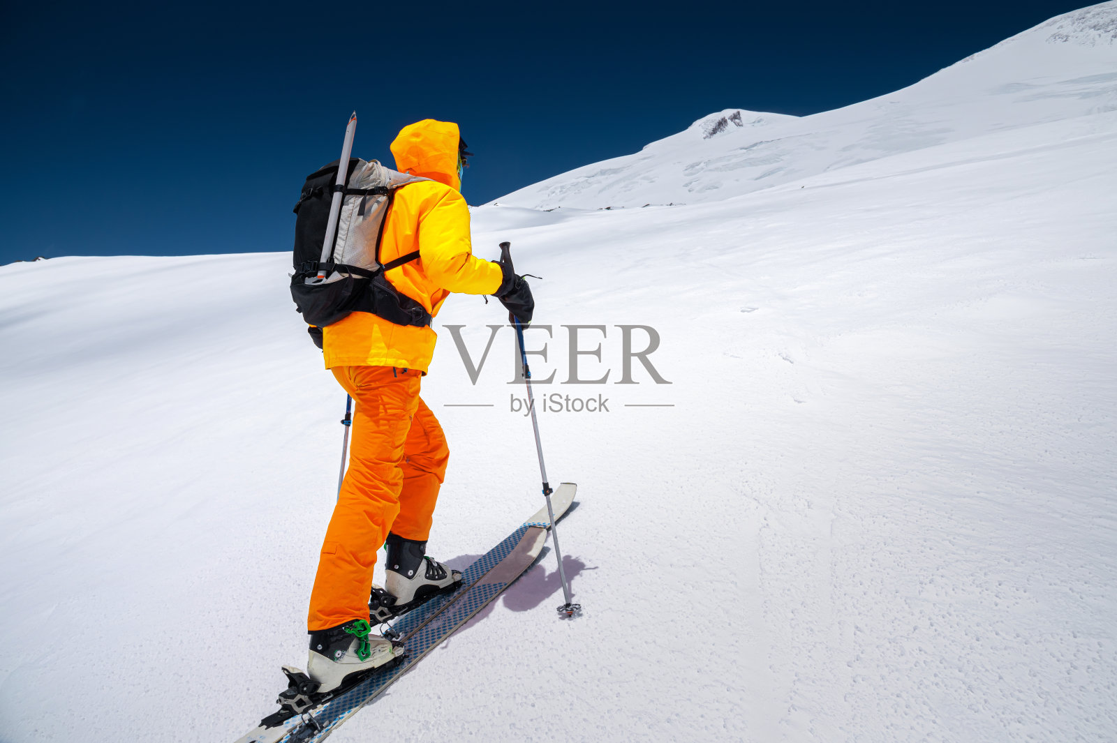 一个阳光明媚的日子里，年轻的男性自由骑手向导穿着黄色的滑雪服，肩上背着背包，骑着滑雪板爬山。照片摄影图片