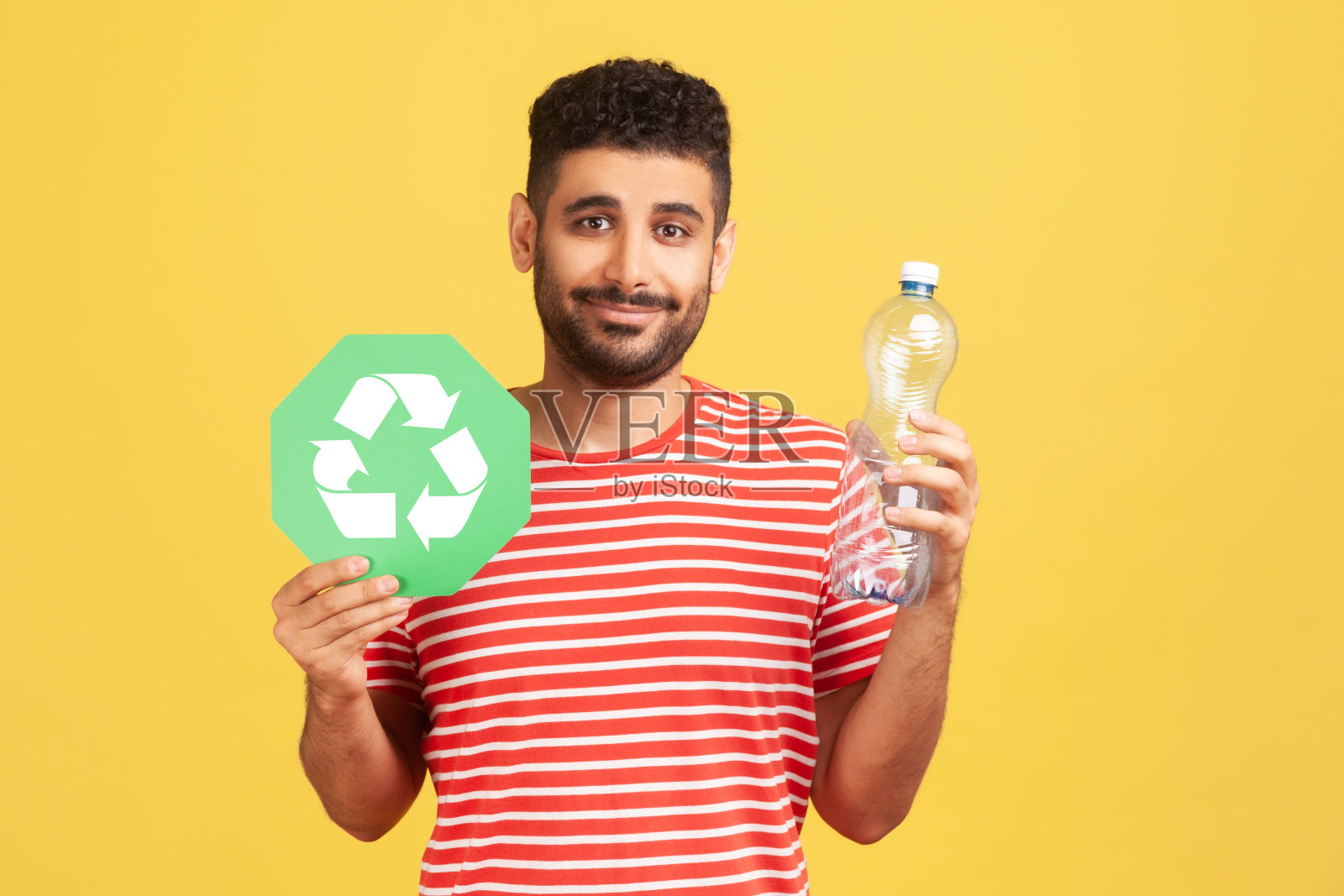 满脸笑容的正面男子，胡子条纹t恤，拿着塑料瓶和回收绿色标志，分类他的垃圾，拯救生态照片摄影图片
