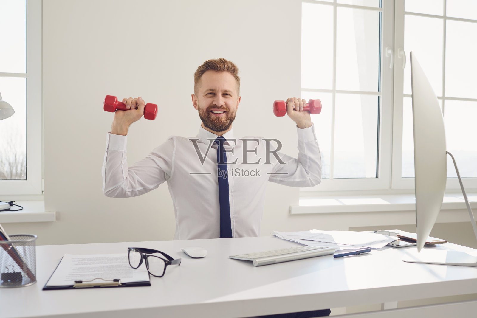 快乐的成功商人穿着白衬衫在办公室里举着哑铃锻炼。照片摄影图片